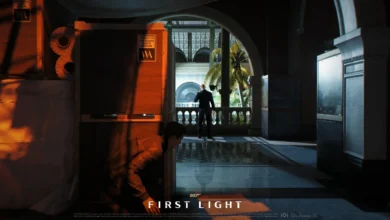 A person crouches behind shipping crates in a dimly lit museum, observing a man in a tuxedo who stands on a balcony overlooking a courtyard.