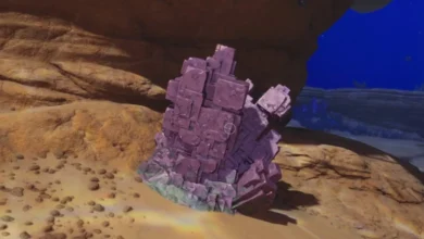 A large cluster of purple, cubic crystals sits on a sandy seabed beneath a rocky overhang.