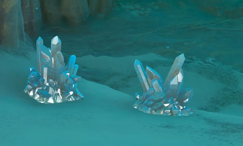 Two clusters of translucent blue crystals resting on the sandy floor of a dimly lit, bluish cavern.