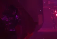 A hooded character hides in the shadows, observing a glowing robotic guard in a futuristic, red-lit environment.