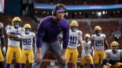 An LSU football coach wearing a headset and purple gear stands focused on the sideline with players in uniform behind him in a stadium.